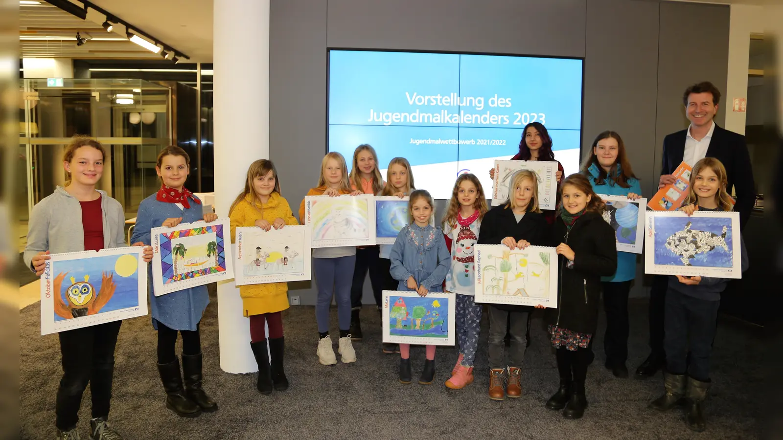 Stolz präsentieren die Kinder und Jugendlichen den Jugendmalkalender 2023 mit Bankvorstand Klaus Berger. (Foto: VR Bank)